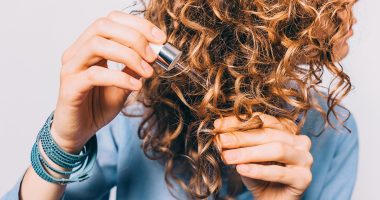Young woman in blue shirt holding her curly hair tips and pipette with oil. Female's hands applying nutritional serum on split ends.